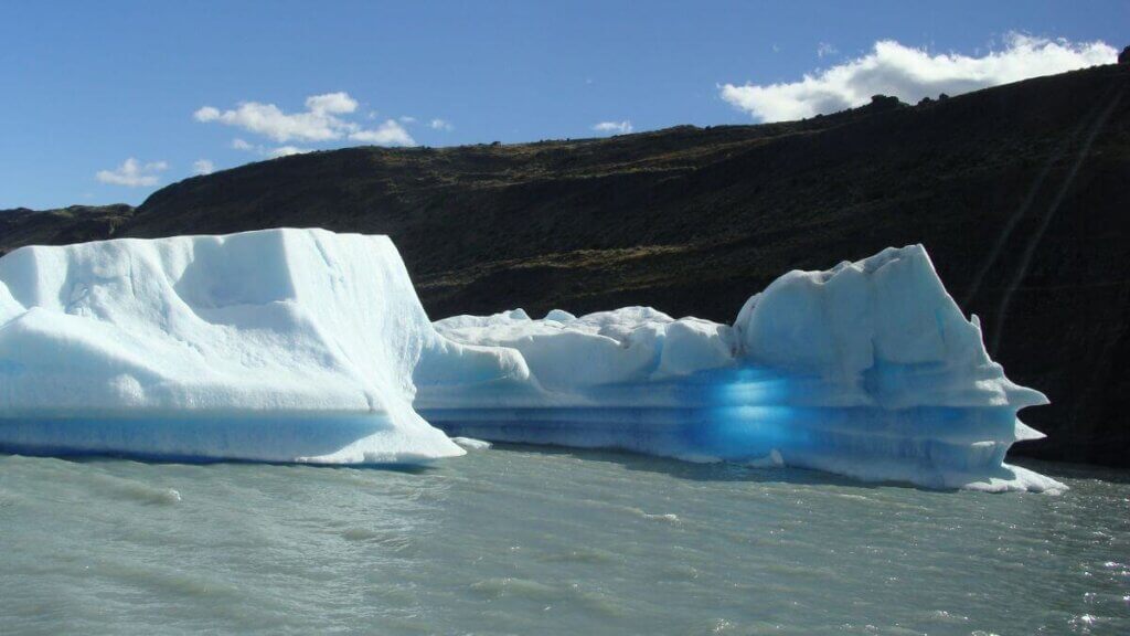 Grande bloco de gelo com formato interessante flutuando no Lago Argentino, refletindo tons de branco e azul conforme o sol incide sobre sua superfície.