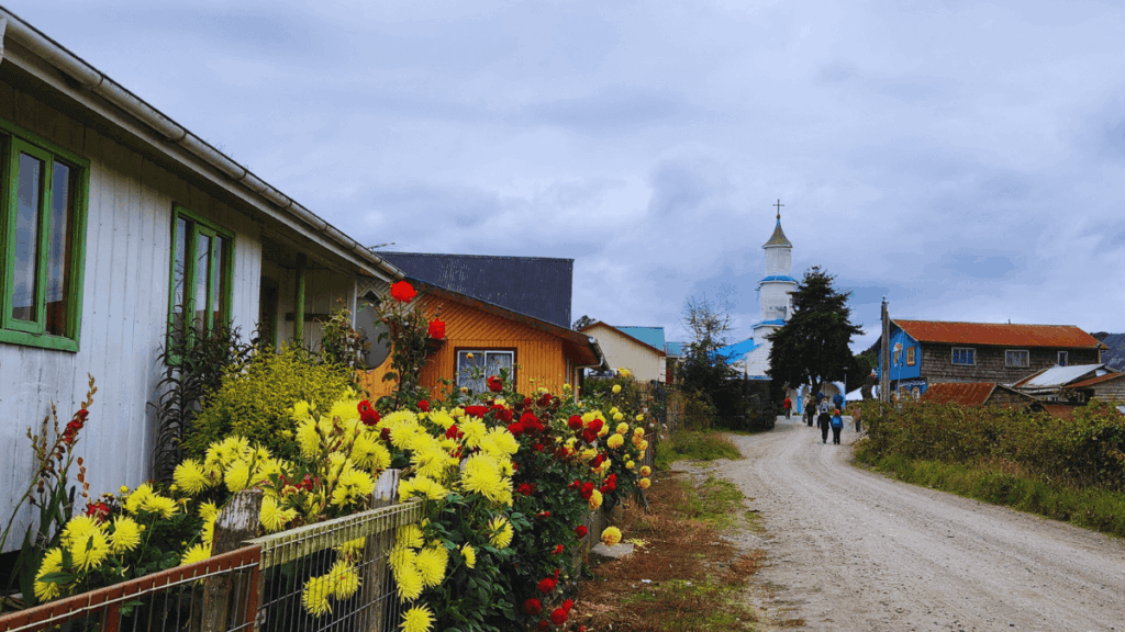 Rua de terra com flores silvestres à beira do caminho e a icônica Igreja de Rilán ao fundo, retratando a paisagem rural autêntica de Chiloé onde natureza e patrimônio cultural se integram harmoniosamente.