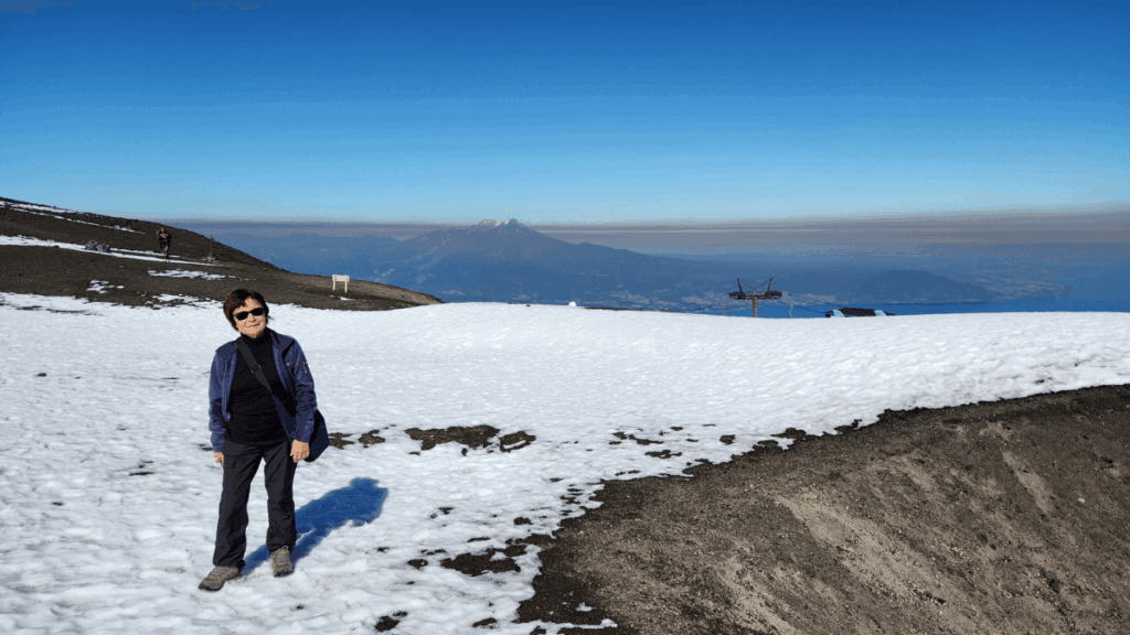 Vista da estação de esqui do Vulcão Osorno em dia de sol, com neve nas encostas, o Lago Llanquihue à direita e o Vulcão Calbuco ao fundo à esquerda.
