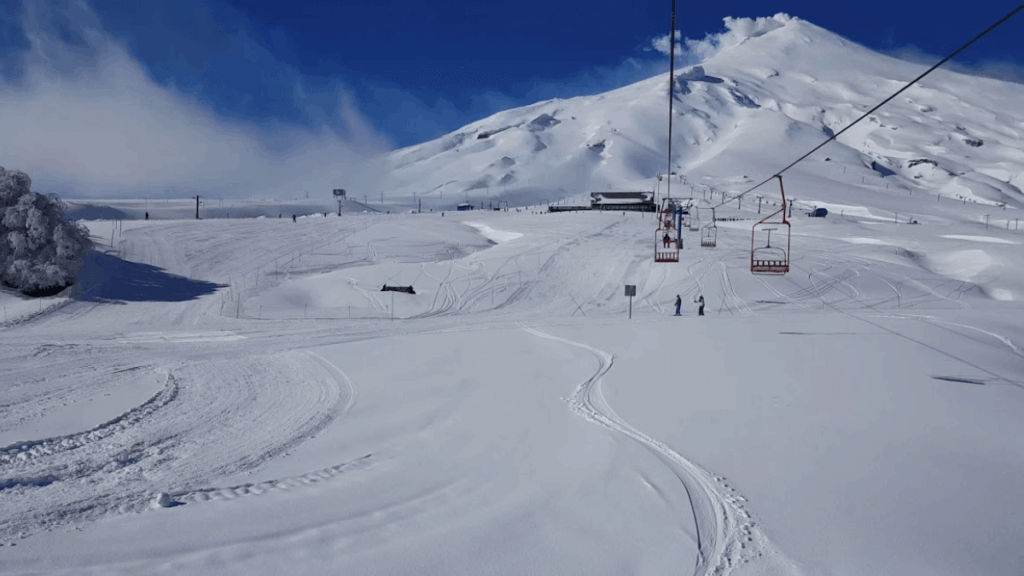 Teleférico da estação de esqui do Vulcão Villarrica em Pucón, com neve e fumaça saindo da cratera em dia claro de inverno.